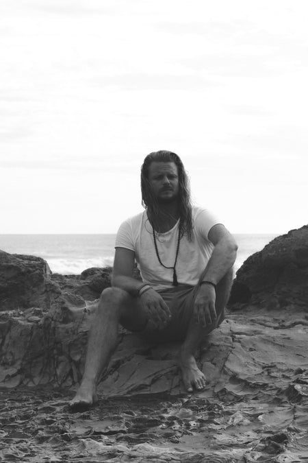 Longhaired man sitting on rocks in front of the sea. Wearing a white t-shirt and a black beaded crystal  necklace