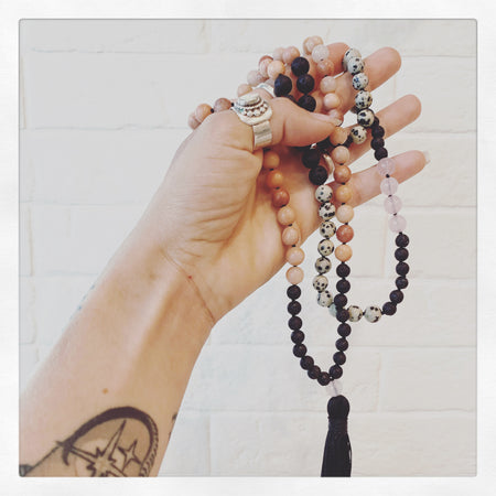 Hand with silver rings holding black lava, rose quartz and rhodochrosite crystal beaded mala necklace with black tassel